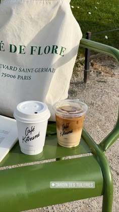 two cups of coffee sit on a green bench next to a book and paper bag