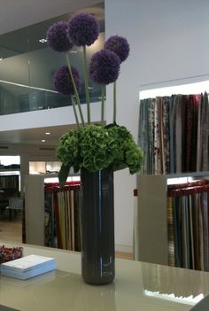 three purple flowers in a vase on a counter with bookshelf in the background