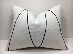 a white pillow with black piping on the front and back sides, sitting against a wall