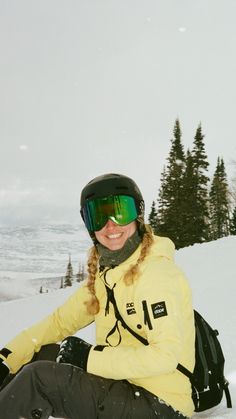 a woman sitting in the snow with her skis on, wearing a yellow jacket and green goggles