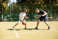 two young men playing basketball on an outdoor court in the sun, with trees behind them