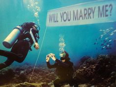 two people in scuba gear standing under a sign that says will you marry me?