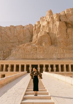 a woman is standing on some steps in front of a mountain and rock formation with her arms outstretched