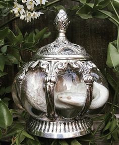 an ornate silver teapot sitting on top of a wooden table surrounded by greenery