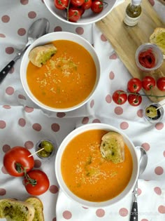 two bowls of tomato soup on a polka dot tablecloth with bread and tomatoes in the background