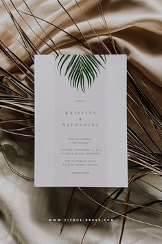 a wedding card with palm leaves on it sitting on a satin material background that is soft and elegant