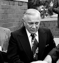 an older man in a suit and tie sitting at a table with other people around him