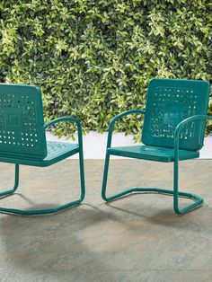 two green chairs sitting next to each other near a wall with bushes in the background