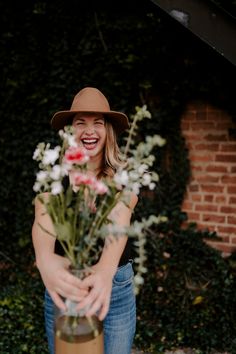 a woman wearing a hat holding a vase with flowers in it and smiling at the camera