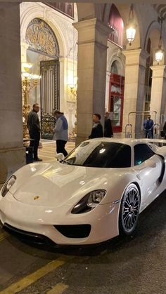 a white sports car parked on the street in front of a building with people standing around it