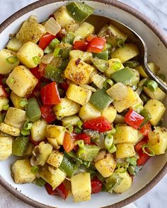 a white bowl filled with potatoes and peppers on top of a marble countertop next to a metal spoon