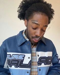 a man wearing a blue leather jacket looking at his cell phone while standing in front of a white wall