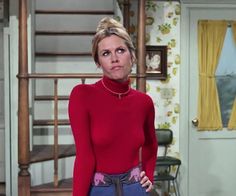 a woman in a red shirt and blue pants is looking at the camera while wearing a necklace