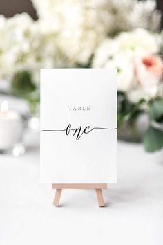 a place card holder with the word table one written on it and flowers in the background