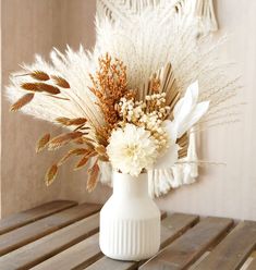 a white vase filled with lots of flowers on top of a wooden table next to a wall