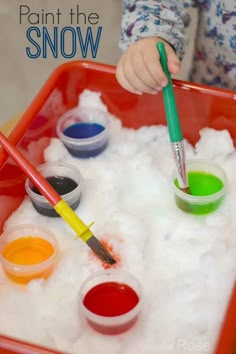 a child is painting in the snow with paintbrushes and watercolors on a red tray