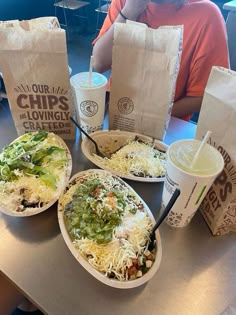 three burritos and two drinks on a table with paper bags in front of them