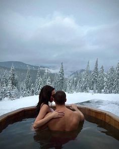 a man and woman in a hot tub with snow on the ground behind them, surrounded by trees