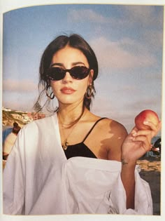 a woman wearing sunglasses holding an apple in her right hand and looking at the camera