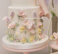 a white cake with pink and yellow flowers on the top is surrounded by cupcakes