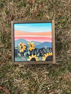 a painting is laying in the grass with flowers painted on it's side and mountains in the background