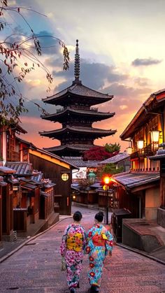 two people are walking down the street in front of some pagodas and buildings at sunset