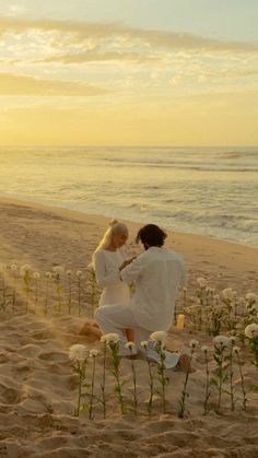 two women in white dresses standing on the beach with flowers and water behind them at sunset