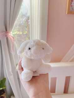 a hand holding a small white stuffed animal in front of a window with pink curtains