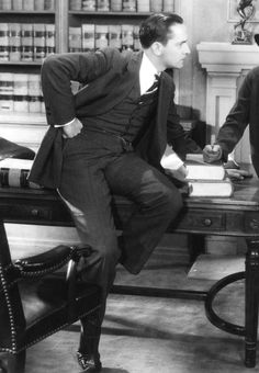 two men in suits sit at a desk with books on it and one is holding his head