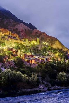 the village is lit up at night on the mountain side with trees and bushes in front of it