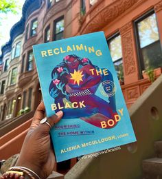 a person holding up a book about reclaiming the black body in front of some buildings