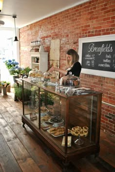 a woman standing behind a counter filled with pastries and doughnuts in front of a brick wall