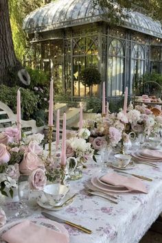 a table set with pink flowers and candles for a formal dinner in a garden setting