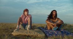 two people sitting on a blanket in the middle of an open field, one holding a book