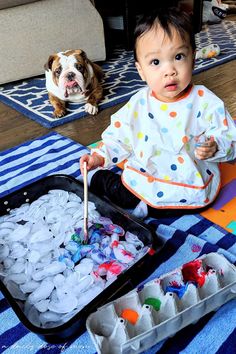 a baby sitting on the floor next to an open tray of ice cubes and a dog