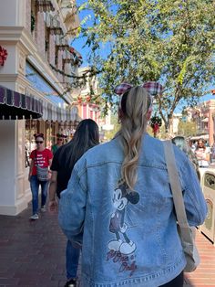 a woman walking down a street with a mickey mouse denim jacket on and back to the camera