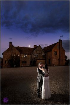 a bride and groom standing in front of a large house at night with the moon behind them