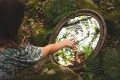 a woman pointing at a circular mirror in the middle of some moss covered rocks and trees