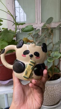 a hand holding a ceramic panda bear figurine in front of a potted plant