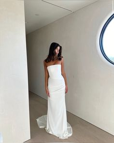 a woman in a white dress standing next to a window and looking at the camera