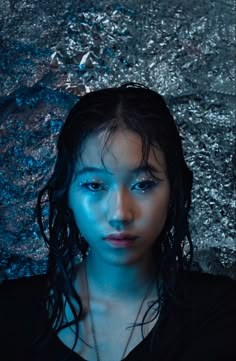 a woman with blue makeup and long hair standing in front of a stone wall, looking at the camera