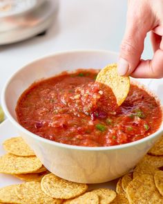 a hand dipping a tortilla chip into a bowl of salsa with tortilla chips on the side