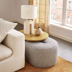 a living room with a round ottoman and lamp on the end table in front of a window