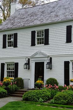 a large white house with black shutters and flowers in the front yard on a cloudy day