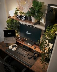 a desk with two computers on top of it next to plants and a clock in the background