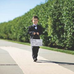 a young boy in a tuxedo is walking down the street holding a sign