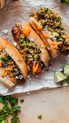 three hot dogs with toppings sitting on top of paper next to lime wedges