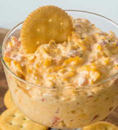 a glass bowl filled with dip and crackers on top of a wooden table next to some crackers