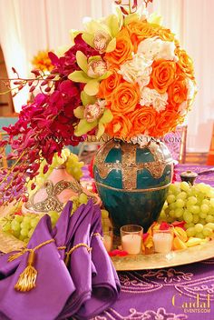 a table topped with a vase filled with flowers next to grapes and oranges on top of a plate