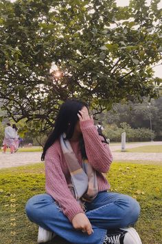 a woman sitting on the ground talking on her cell phone in front of a tree